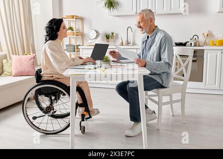 A man and a woman in wheelchair sit together at a table in their kitchen at home. Stock Photo