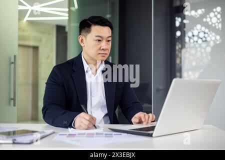 A professional businessman is immersed in work on his laptop, sitting at a sleek table in a contemporary office setting, exemplifying focus and determination. Stock Photo