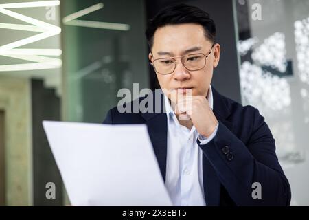 A middle-aged Asian businessman deeply focused, examining documents with a thoughtful expression, in a contemporary office setting with ambient lighting. Stock Photo