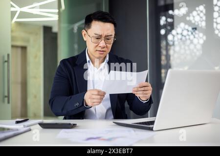 Focused businessman in a dark suit studying a paper document while sitting at a sleek, contemporary office desk with laptop and smartphone. Stock Photo