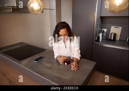 View from above of a young pretty woman in white bathrobe, making tea for the breakfast. Charming young woman stirring sugar in a cup of tea while hav Stock Photo