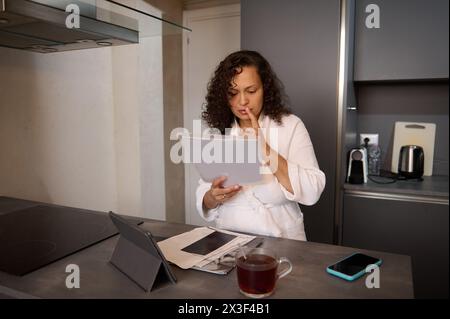 Young serious woman in white bathrobe, analyzing documents, doing paper work at home. Multi ethnic pretty brunette holding hands on lips while going t Stock Photo