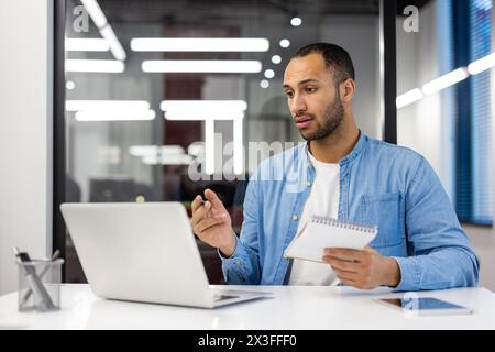 A young Latin American male student studies remotely, sits in the office at a desk, records a lecture, communicates via video call on a laptop. Stock Photo