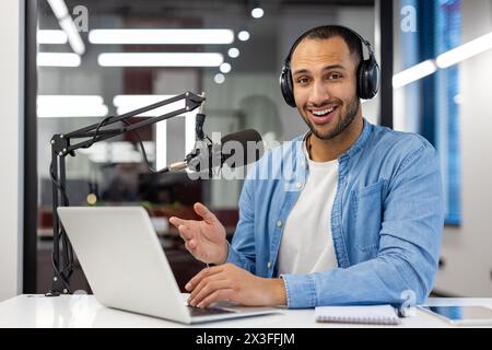 Portrait of a smiling young Muslim man sitting in headphones in the office at the table, talking online through a microphone, looking at the camera. Stock Photo