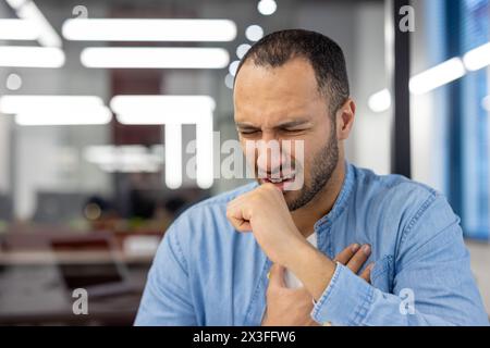 Close-up photo of a young sick hispanic man sitting in the office and coughing covering his mouth with his hand and holding his chest. Stock Photo