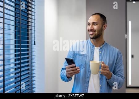 A smiling and relaxed young Muslim man rests in the office, stands in the corridor of the office by the window, holds a phone and a cup of coffee in his hands, looks to the side. Stock Photo