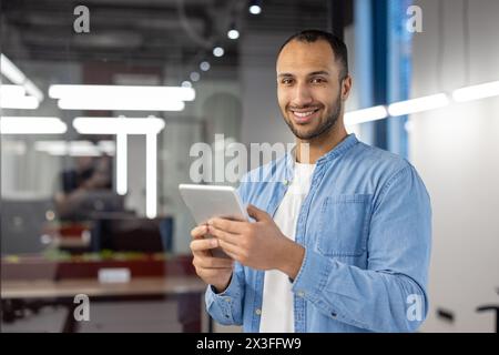 Portrait of a young hispanic engineer engineer standing in a modern office, holding a tablet and smiling at the camera. Stock Photo