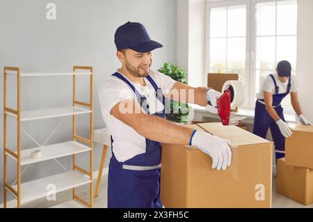 Young skilled male worker of moving service glues cardboard box with duct tape. Stock Photo