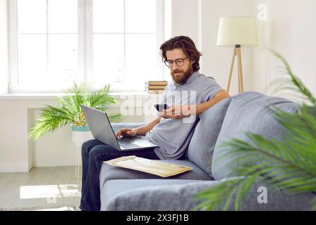 Bearded Caucasian man with laptop on knees sits on sofa with postal envelope and uses phone Stock Photo