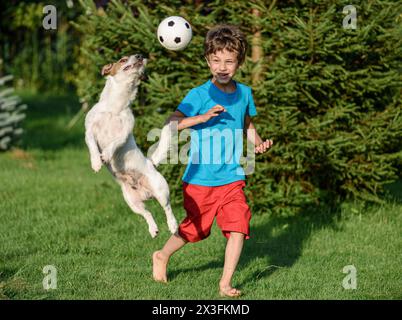 Kid playing football with family pet dog in backyard garden. Dog jumps to catch a ball. Stock Photo