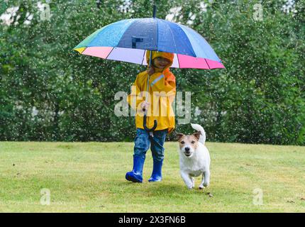 Kid in raincoat under umbrella and her dog playing on grass lawn during spring rain Stock Photo