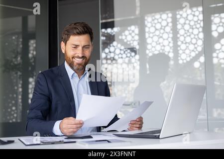 Portrait of a smiling young businessman sitting at an office desk with a laptop and looking through documents and bills. Stock Photo