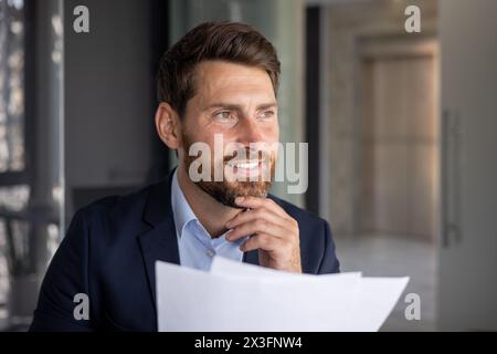 Close-up photo of a young businessman sitting in the office in a suit, holding documents in his hands, thoughtfully and smilingly looking to the side. Stock Photo