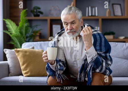 Sad senior gray-haired man sitting on sofa at home and suffering from illness. Covered with a blanket, holding a cup of hot drink and a napkin. Stock Photo