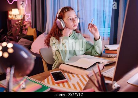 Medium shot of modern teen girl wearing headphones working on school project in her room at home in evening Stock Photo