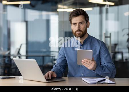 Portrait of a serious young male developer and engineer sitting in the office at the table and working on a laptop and a tablet. Looks confidently into the camera. Stock Photo