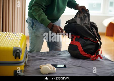 Hands of young unrecognizable man in casualwear bending over bed and putting passports in backpack while preparing for vacation Stock Photo