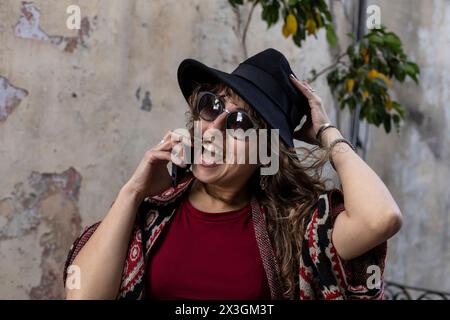 Young Latin American woman with curly hair, hat, sunglasses, positive attitude talks on cell phone Stock Photo