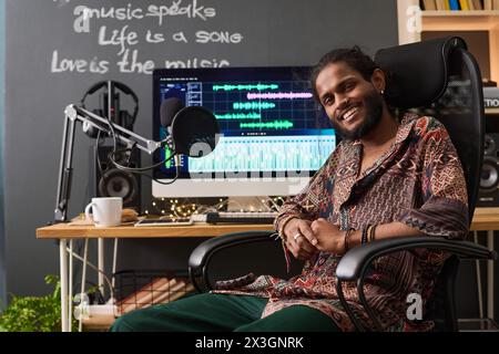 Young successful male artist in ethnic attire looking at camera with smile while sitting by workplace with sound recording equipment Stock Photo