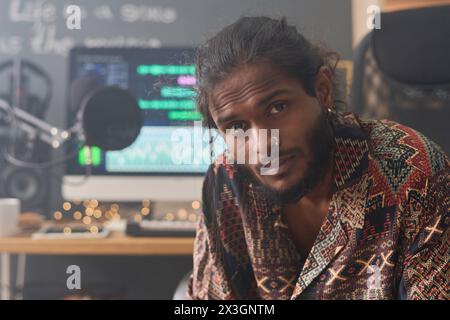 Young creative man with wrinkled forehead sitting in front of camera by workplace with computer monitor and looking at you Stock Photo