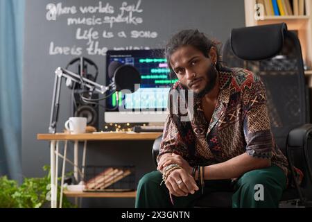 Young confident artist looking at camera while sitting in armchair by his workplace with sound recording equipment in home studio Stock Photo