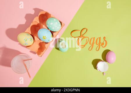 Some lovely Easter eggs contained inside an egg carton painted in coral color. A glass cup filled with water containing two paintbrushes. The word Egg Stock Photo