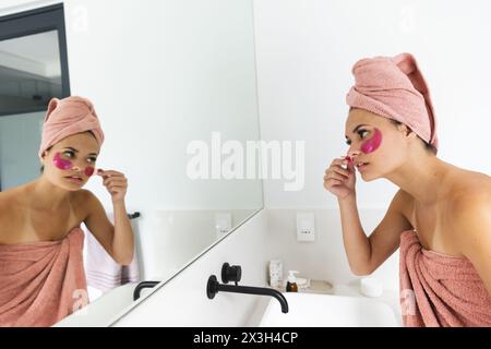 A young Caucasian woman wearing towel, applying a face mask in a bathroom Stock Photo