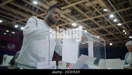 Close up shot of polling box standing on table in polling station. Multi ethnic American voters come and put bulletins in box. Political races of US presidential candidates. National Election Day. Stock Photo