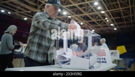 Close up shot of polling box standing on table in polling station. Elderly voter puts bulletin into box and starts look at camera. Political races of US presidential candidates. National Election Day. Stock Photo