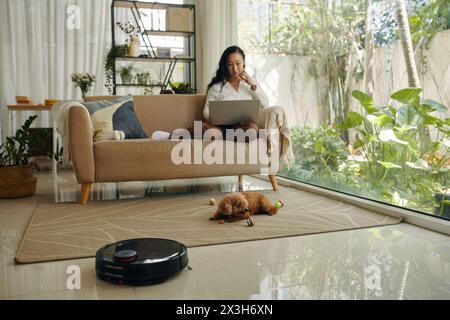 Robot vacuum cleaning floor when young woman working on laptop and dog playing with toys Stock Photo