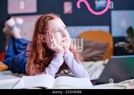 Portrait of modern teen girl with curly read hair daydreaming while doing homework in her bedroom, copy space Stock Photo
