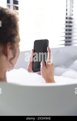 A young Caucasian woman relaxing in bathtub at home, holding a smartphone, copy space Stock Photo