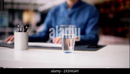 Glass Of Water On Desk And Man In Background Using Computer Stock Photo