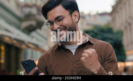 Close up happy lucky gen z Indian Arabian ethnic man guy male businessman student winner holding mobile phone pointing finger fist gesture enjoy Stock Photo