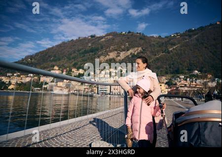 Beautiful smiling woman and her little kid, cute daughter walking on the promenade near the lake of Como. Stylish baby pram in the foreground. Family. Stock Photo