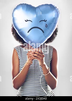 Black woman, emoji and sad heart balloon in studio for heartbreak, depression or emotional on isolated white background. Icon, tear and female model Stock Photo