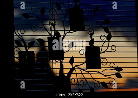 Silhouette of a decorative metal tree with leaves and frames against a sunset through blinds. Stock Photo