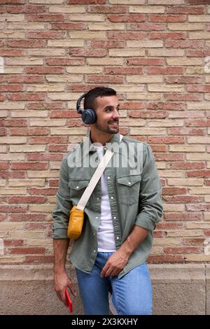 Young stylish man looking smiling to side with wireless headphones holding cell leans brick wall. Stock Photo
