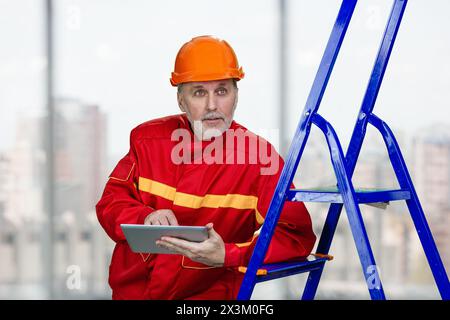 Male elder contruction worker with tablet pc. Aged man with red helmet leans on a blue ladder. Indoor window background. Stock Photo