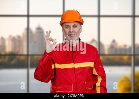 Happy smiling construction worker in red uniform shows okay gesture sign. Checkered window background. Stock Photo