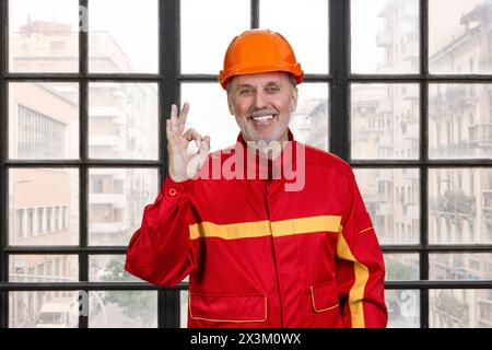 Smiling construction handyman worker in red uniform shows okay gesture sign. Checkered window background. Stock Photo