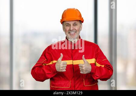Portrait of a smiling handyman construction worker showing both thumb up. Blurred windows background. Stock Photo