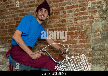 Young Latino male (28) riding a bicycle with basket on his way to work. Copy space, brick background. Concept Jobs Stock Photo