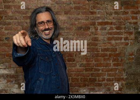 Latin American man (44) with long gray hair, blue jeans jacket points with one arm outstretched in front while smiling. Copy space. Brick background. Stock Photo
