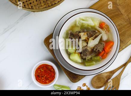 Sop daging sapi or Indonesian beef soup, served on ceramic white bowl with sambal and slices of lime. Selective focus with white marble background. Stock Photo