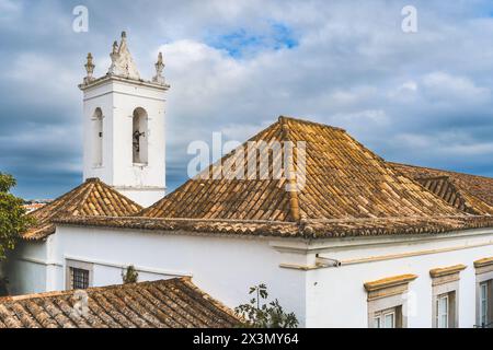 A rooftop view in Tavira, Portugal, showing traditional terracotta roofs, a church tower, and a satellite dish under a cloudy sky. Stock Photo