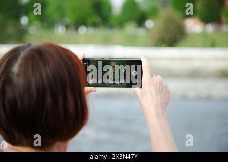 Caucasian woman taking a video with a cell phone camera, standing on the river bank at sunny summer day . Video creator concept Stock Photo