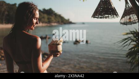 A woman standing on a beach in Thailand, holding a coconut cocktail drink in her hand. Stock Photo