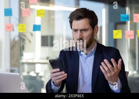 Sad disappointed businessman reading news on phone, man in business suit using app on smartphone, looking upset, received bad news online in email, working with laptop inside office. Stock Photo