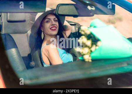 A woman is driving a car with a bouquet of flowers in the back seat. She is wearing a hat and has her eyes closed. Stock Photo